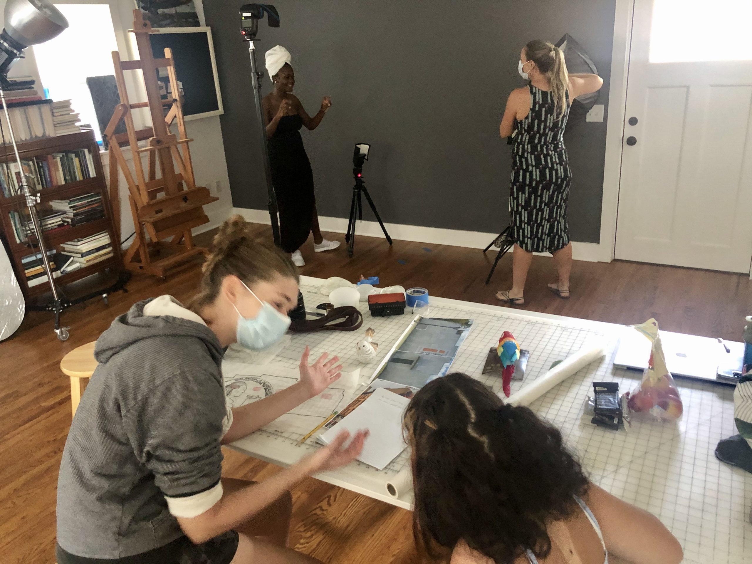 Two women looking at designs on drafting table with two women in the background with photography equipment
