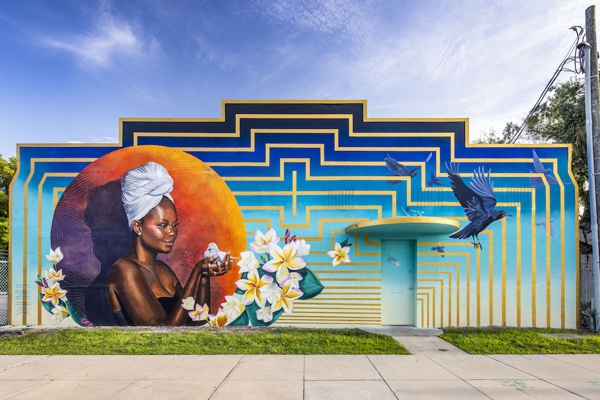 Colorful outdoor mural of Black woman wearing a white head wrap holding a white dove within a circle; image includes ravens and plumeria