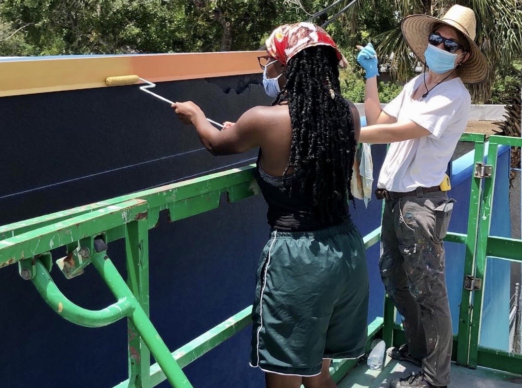 A woman with a scarf around her hair and also wearing a mask rolls orange paint on to a mural from a riser. A second woman who is working is also wearing a mask and gloves and large sunhat. Two women painting a mural from a rise. Both are wearing masks and