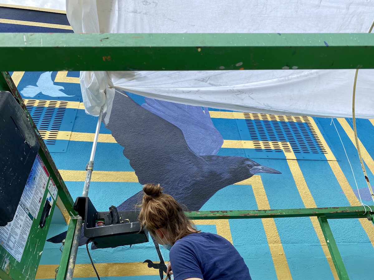 woman painting mural that depicts a raven