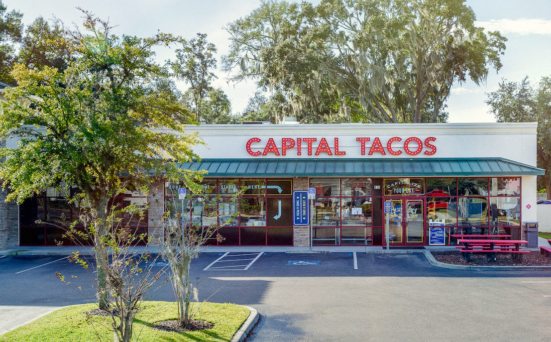 White building with large red letters, Capital Tacos. Red picnic table in front as well as parking.