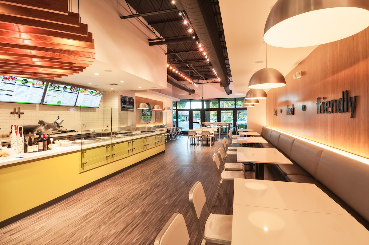 Interior of quick service restaurant. White tables, white chairs, padded banquettes, yellow order counter with menu board.